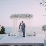 Wedding ceremony beneath a white floral arch overlooking the ocean